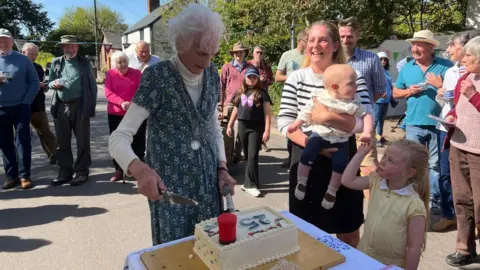 A woman is cutting a cake with a knife outside in the sun while two children are standing next to her.