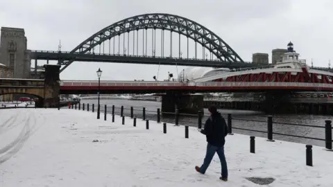 Man in black jumper and black beanie walking on snow covered quayside in front of green Tyne Bridge and red Swing Bridge in Newcastle