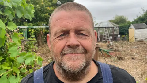 Stephen Jalland is standing in a garden which has two outbuildings on the right and trees on the left. He has short black hair and stubble and is wearing a black T-shirt and overalls.