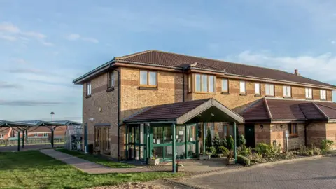 Mike Brown Photographic The outside of Lakelands Hospice, which is a large two-storey brick building with a green porch entrance.