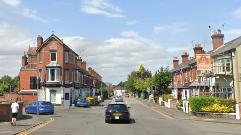 A crossroads with traffic lights on a sunny day. There are terraced brick houses on either side of the street running straight ahead. There is a person in a white t-shirt and black shorts walking along the street.