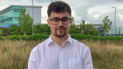 Alex Whinfrey-Gibson stares at the camera. He has brown hair and a brown beard and is wearing a white, open-necked shirt and dark framed glasses