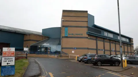 A large prison building, with a car park next to it. The building is sandstone coloured, with a sign saying Barlinnie in large letters on the wall