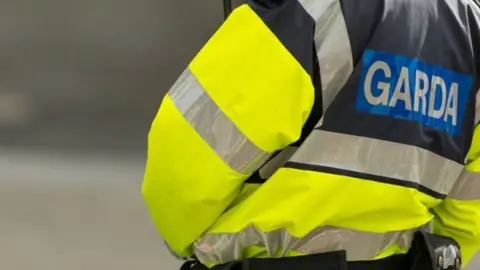 A person in a yellow garda jacket, with blue garda sign on the back.
