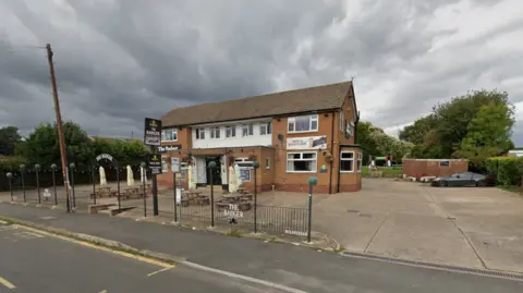 Google A pub with seating out the front, with grey skies above it. There is an empty car park outside and the road can be seen in the foreground, with a telegraph pole to the left of the shot.