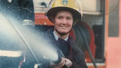 An old image of a female firefighter holding a hosepipe and spraying water. She wears a yellow helmet and a large black protective jacket.