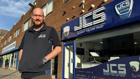 A man with thin-rimmed glasses and a beard wears a blue gilet and stands in front of his shop, which has a logo formed from the initials JCS.