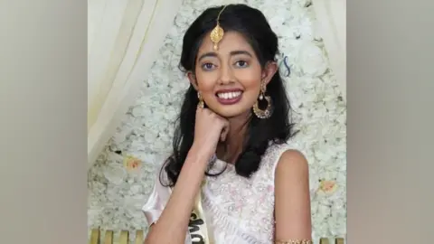 Family Sudiksha Thirumalesh smiling in a white dress with an embroidered floral design and wearing gold jewellery 