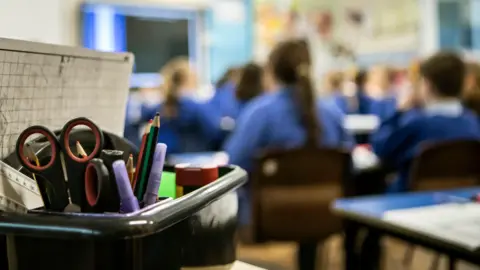 PA Media A generic image of a classroom with children in the midground in blue jumpers. There is a board on the wall showing teaching aids and there is a box of pens and pencils in the foreground.