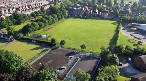 An aerial view of a large square playing field surrounded by trees and hedges with rows of houses nearby.