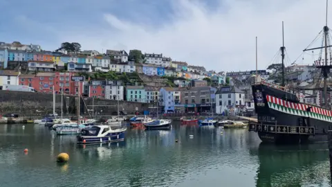 A number of small boats sit idly in Brixham harbour against a backdrop of small, colourful houses 