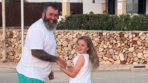 Family A girl wearing a white sleeveless top is standing next to, and holding hands with, a man wearing a white t-shirt. He has a beard and tattoos. They are standing outdoors with a wall and hedge in the distance.