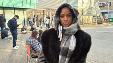 BBC/Simon Jones Young woman wearing hood, scarf and coat with others queuing outside behind her outdoors