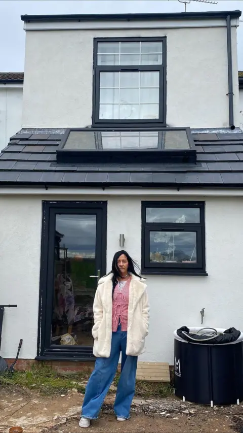 Suzie Cavadino, who has long, black hair, wears blue jeans and a white coat over a red-and-white gingham top. She is standing in front of the narrow, two-storey extension that occupies the back of her house.