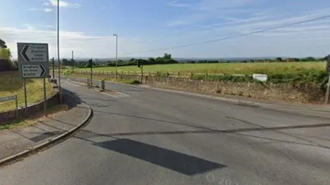 Google Leigh Crossroads as seen on a sunny day with road signs and a low stone border wall on a sunny day with blue skies overhead