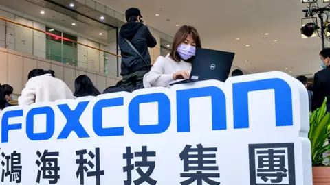 Getty Images A woman wearing a face mask seen working on her computer on top of Foxconn's logo