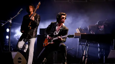 Frontman Alex Turner plays guitar on stage. He has brown hair and wears sunglasses and black blazer over a white shirt. The band's guitarist and pianist are in the background. 