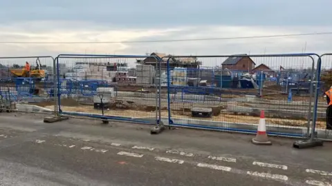 Andrew Turner/BBC Fencing surrounds a works site where a central reservation and pedestrian crossing is being built. In the background is machinery and houses under construction.