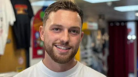 Sion Williams, with light beard, wearing grey jumper and beige baseball hat in front of clothing rack in his shop.