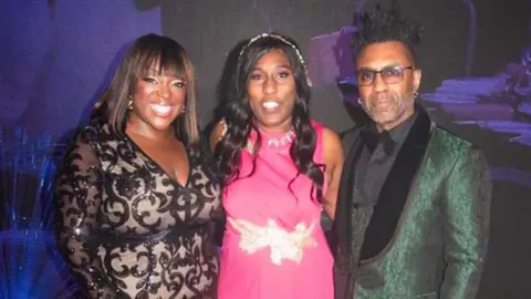 MBCC Two women and a man stood against a purple backdrop looking at the camera. The woman on the left is wearing a black and beige dress and the woman in the middle is wearing a pink dress. The man, stood on the right, is wearing a green tuxedo