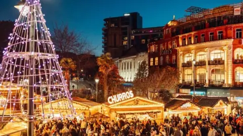 Bournemouth Christmas Market Crowds around lit-up market stalls with a large wire Christmas tree and buildings around the edge.