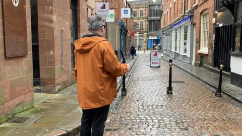 A man wearing an orange coat and holding a white stick looks away from the camera, down a cobbled street lined with shops and pubs.