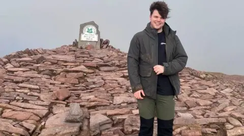 Jordan Digby Jordan standing at the summit of Pen Y Fan in a coat