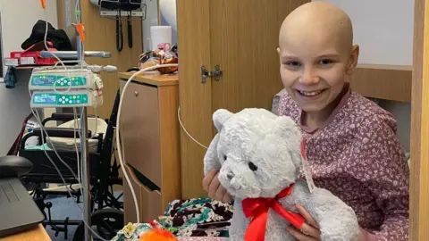 Ian and Sam Frisby Poppi in a hospital bed cuddling a large teddy bear. She is wearing red pyjamas with a white pattern and is smiling. She has lost her hair because of her treatment. There is a medical machine and a chest of drawers next to the bed.