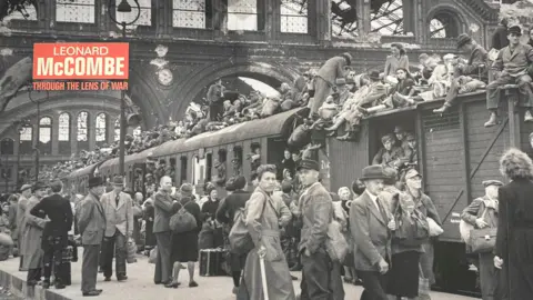 LEONARD MCCOMBE Train carriages at a railway station filled with people inside on sitting on top, with many others on the platform in front of it.