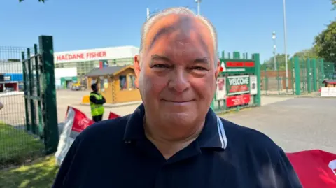 A man wearing a navy polo top. He has short grey hair and is looking at the camera.
