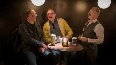 Channel 4 Producer Phil Collinson, writer Russell T Davies and actor Alan Cumming are standing around a table and laughing while on set for episode five