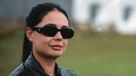Hannah Laing, in dark sunglasses, her dark hair in a ponytail and wearing a black leather jacket, stands with a large "Doof" sign behind her in Camperdown Park
