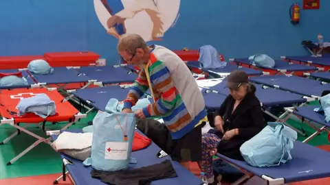 A man and woman at the centre of the frame with temporary bunk beds have been set up in an evacuation centre.
