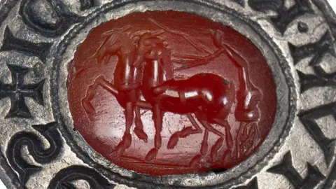 A close-up of an oval silver seal matrix, with a hanging loop on the left side. It is resting on its side, to show the carving of two horses, with upraised legs and a charioteer leaning over them from his chariot with a whip on the oval red gemstone intaglio in the centre. Its thick silver rim is carved with letters. 