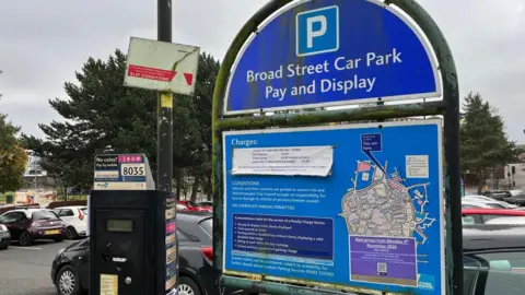 A blue notice board in a car park with the words Broad Street car park pay and display. Cars can be seen parked in lines behind it and a pay and display machine on the left of the sign.