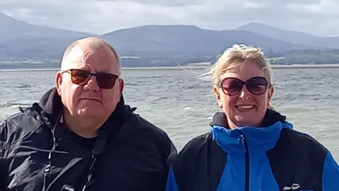 Norman and Amanda Barber, both wearing waterproof blue coats and sunglasses, smile for the camera with the sea behind them