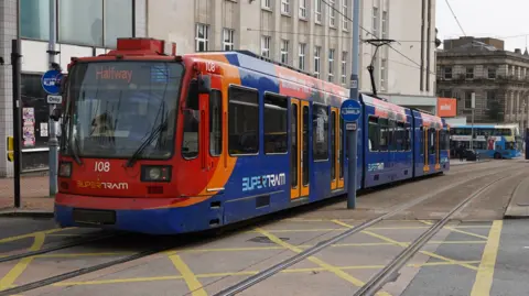 Oli Constable/BBC A red and blue coloured tram runs through Sheffield city centre. Its destination is marked 'Halfway'