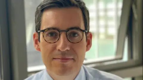 A man with dark hair and oval glasses looks directly at the camera, smiling