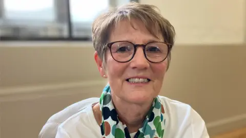 Alex Dunlop/BBC Angela Ellis, sitting down, looking at the camera and smiling, wearing glasses with short fair hair. She has a scarf round her neck and a white top on. She is in a room with cream walls behind her. 