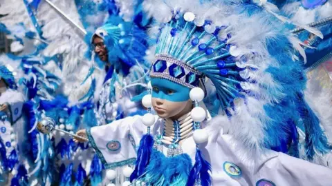 A very striking image of a child dressed in a blue and white feathered outfit, with a large blue and white feathered headdress and blue paint across the top part of their face. Their arms are outstretched and there are people in the background wearing similar outfits.