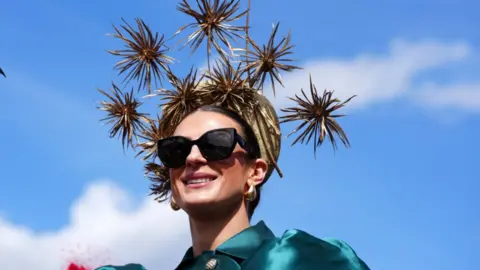 PA Media A woman in a gold hat with thistle style adornments spiking from it. She wears a green vintage style dress and black sunglasses.