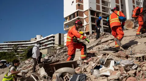 EPA Rescuers in high-vis orange clothing and hard hats walk up a large pile of rubble.