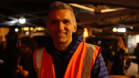 Kian Boyle/BBC A person is standing outdoors at night beneath a canopy, wearing a bright orange reflective safety vest over a thick blue jacket. Around him are other volunteers, some also in reflective gear. 