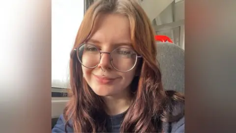 Corinna Baker. She has red hair, is smiling and is wearing a pair of large circular spectacles. She has a nose ring and stud through her lip