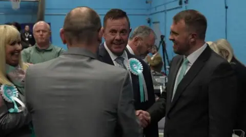 A man in the middle wearing a light blue and white rosette is looking at a man with his back to the camera. Another man on the right is smiling and shaking someone's hand, while a woman with blonde hair is smiling on the far left.
