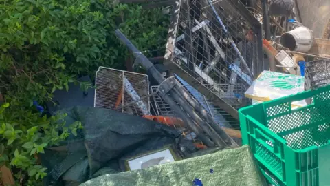 Sandra McCluskey A large pile of dumped waste and scrap materials stacked beside a hedge and a dark vehicle. The debris includes metal fencing panels, poles, crates, tools, tarpaulins and assorted household items, with vegetation partially growing around the rubbish. The scene appears to show fly-tipped waste in an overgrown outdoor area on a sunny day.