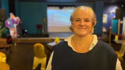 A woman with short blonde hair smiling at the camera wearing a necklace and white shirt and blue sleeveless cardigan. Behind her are tables and chairs and a stage.