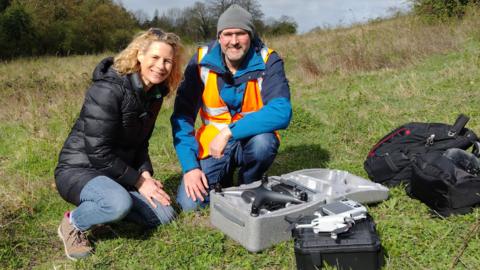Emily Jeffery with drone pilot David Campion. They are in a field, both kneeling on the grass.