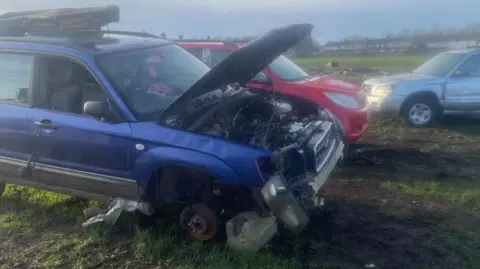 Simon Thake/BBC Three abandoned cars are left on a grass field. The bonnet of the nearest blue car is open with bits of engine spilling out and a large brick in place where the wheel should be.