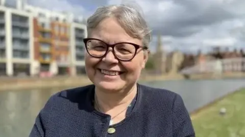 Laura Church with short fair hair, wearing glasses, smiling at the camera and wearing a dark blue buttoned top. She is standing on the bank of a river with a five-storey office block behind.